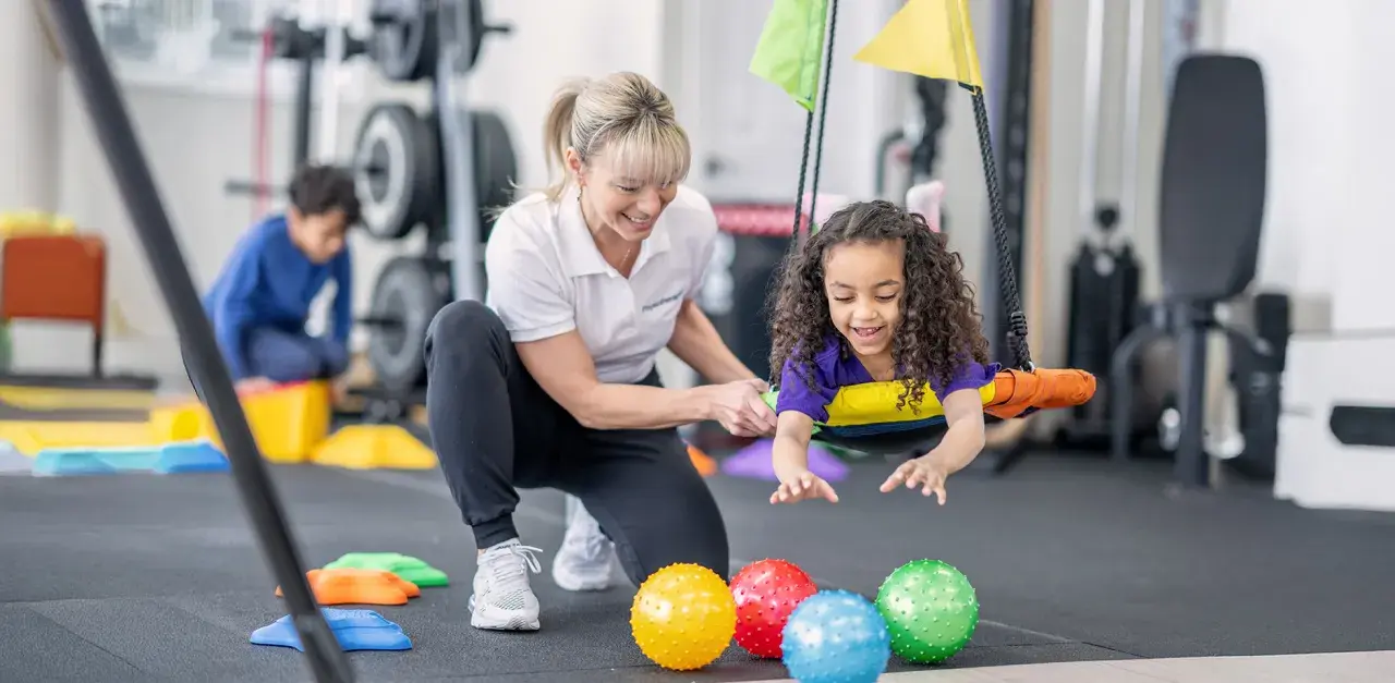 Ergotherapie für Kinder: Ein Mädchen lacht auf einer Schaukel, während eine Therapeutin sie unterstützt. Bunte Bälle liegen im Vordergrund.