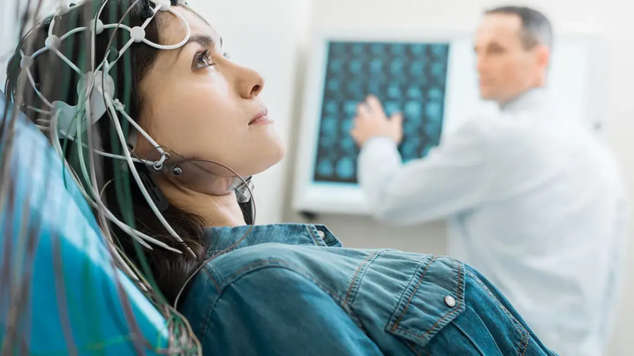 Frau mit EEG-Kappe für medizinische Untersuchung, Arzt im Hintergrund analysiert Gehirnscan.