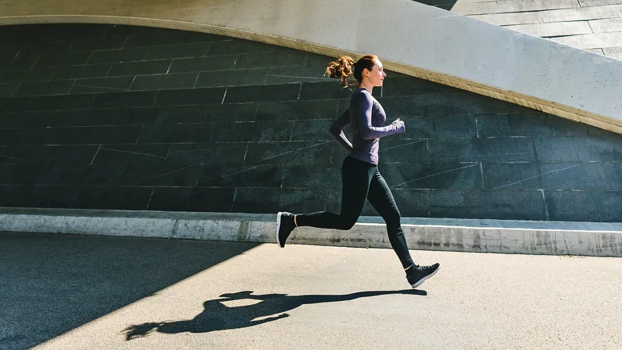Frau im lila Oberteil und schwarzen Leggings beim aerobes Training, ihr Schatten auf dem Asphalt.