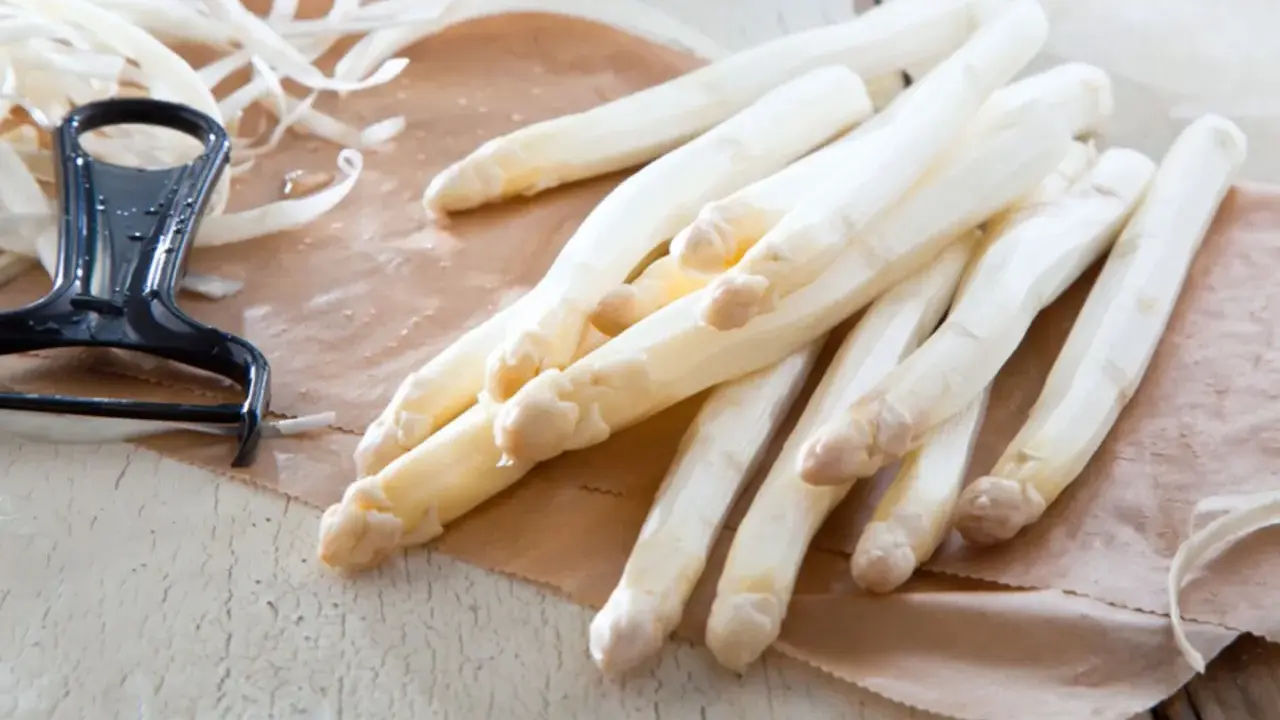 Frischer weißer Spargel liegt auf braunem Papier, daneben ein Sparschäler. Die Frage, warum riecht Urin nach Spargel, beschäftigt viele.