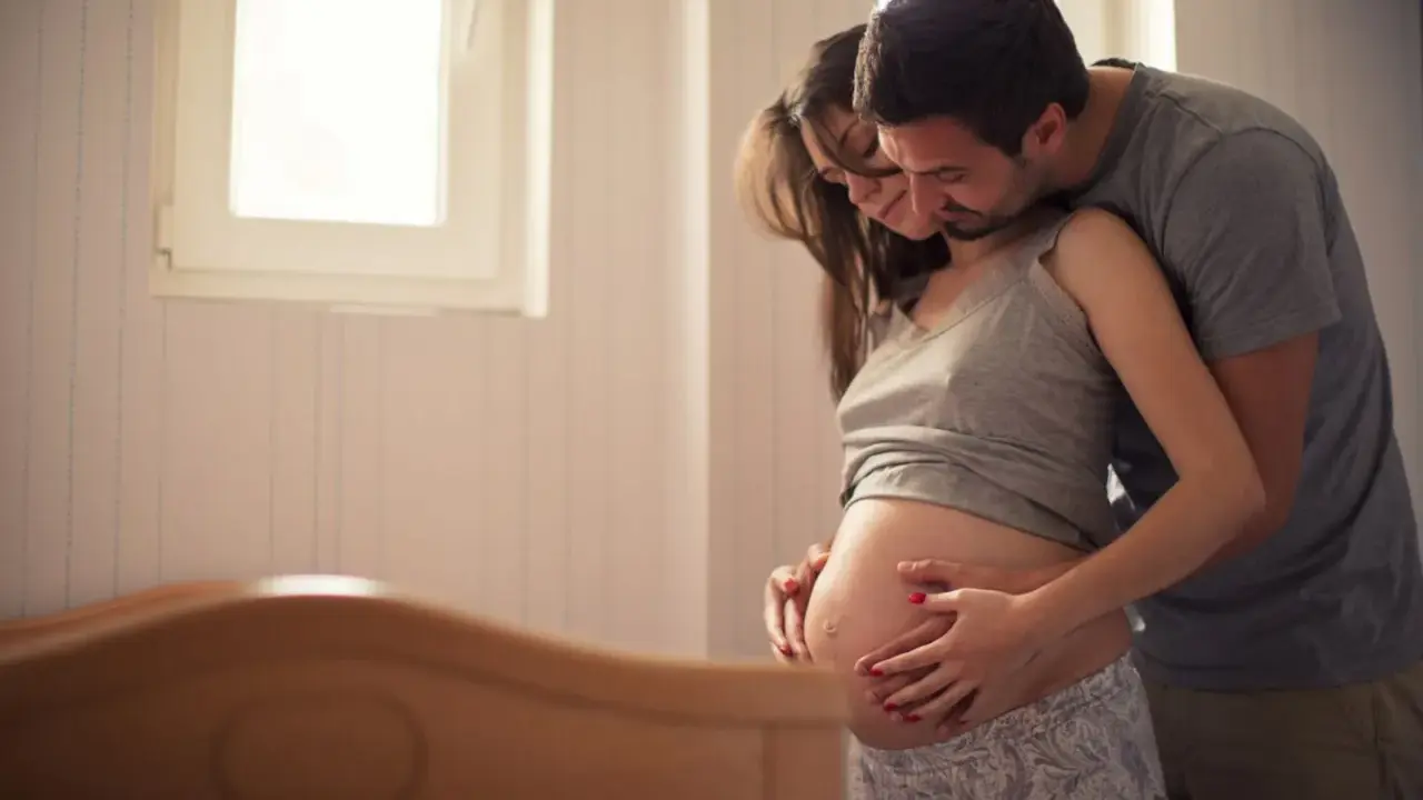 Ein Mann umarmt seine schwangere Frau, die ihren Bauch streichelt. Sie warten gespannt auf ihr Baby. Senkwehen ab wann?