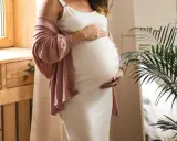 Frau in weißem Kleid und rosafarbener Strickjacke hält ihren Bauch, 17 ssw.