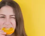 Junge Frau mit einem Stück Orange im Mund, die versucht, nach Weisheitszahn-OP wieder feste Nahrung zu essen.