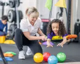 Ergotherapie für Kinder: Ein Mädchen lacht auf einer Schaukel, während eine Therapeutin sie unterstützt. Bunte Bälle liegen im Vordergrund.