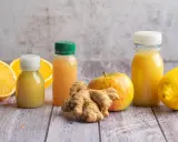 Frische Zutaten für einen belebenden Ingwer-Shot: Orangen, Zitrone, Apfel und Ingwerwurzel neben kleinen Flaschen mit gelbem Saft.