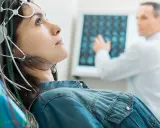 Frau mit EEG-Kappe für medizinische Untersuchung, Arzt im Hintergrund analysiert Gehirnscan.