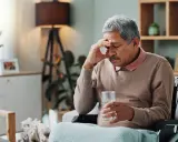 Älterer Mann im Rollstuhl mit Kopfschmerzen, hält ein Glas Wasser. Möglicherweise ein Parkinson-Test der Haut, der auf seine Symptome hinweist.