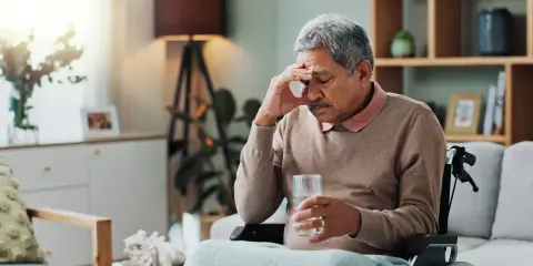 Ein Mann im Rollstuhl hält ein Glas Wasser, die Hand an der Stirn. Ein Bild, das den **Parkinson Verlauf nach 10 Jahren** symbolisiert, mit Müdigkeit und Sorge.