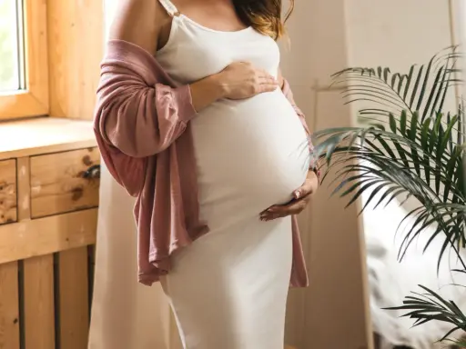 Frau in weißem Kleid und rosafarbener Strickjacke hält ihren Bauch, 17 ssw.