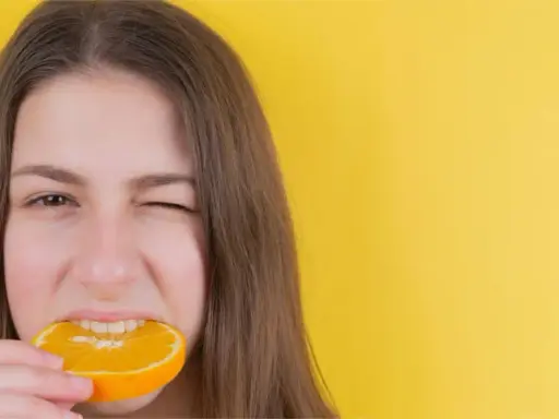 Junge Frau mit einem Stück Orange im Mund, die versucht, nach Weisheitszahn-OP wieder feste Nahrung zu essen.
