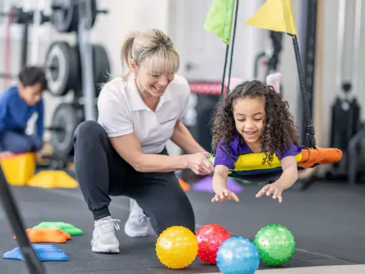 Ergotherapie für Kinder: Ein Mädchen lacht auf einer Schaukel, während eine Therapeutin sie unterstützt. Bunte Bälle liegen im Vordergrund.