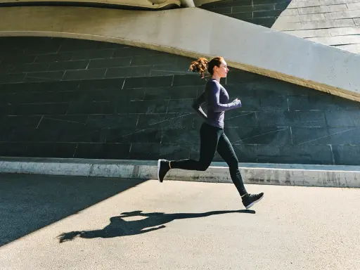 Frau im lila Oberteil und schwarzen Leggings beim aerobes Training, ihr Schatten auf dem Asphalt.