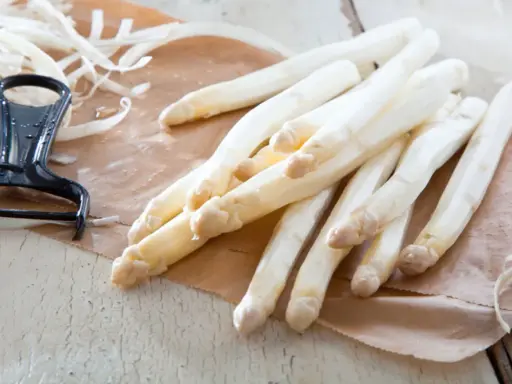 Frischer weißer Spargel liegt auf braunem Papier, daneben ein Sparschäler. Die Frage, warum riecht Urin nach Spargel, beschäftigt viele.