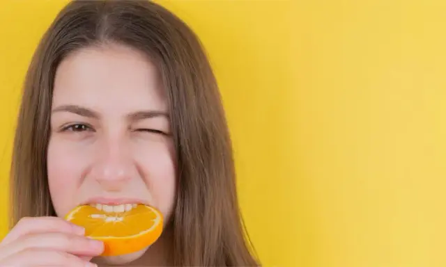 Junge Frau mit einem Stück Orange im Mund, die versucht, nach Weisheitszahn-OP wieder feste Nahrung zu essen.