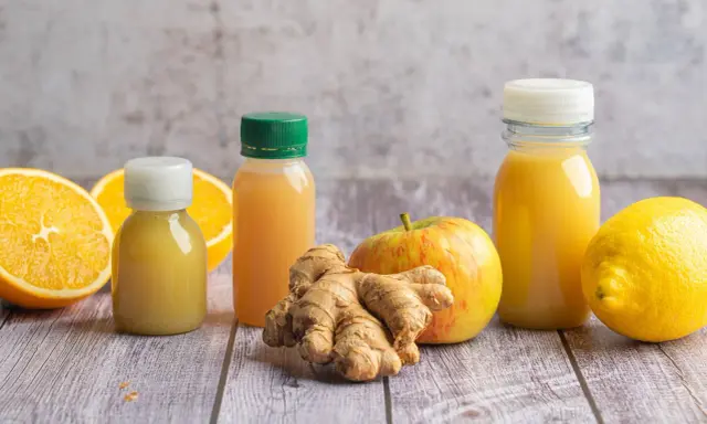 Frische Zutaten für einen belebenden Ingwer-Shot: Orangen, Zitrone, Apfel und Ingwerwurzel neben kleinen Flaschen mit gelbem Saft.