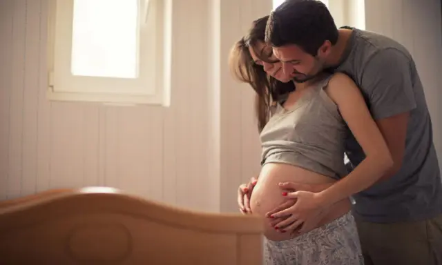 Ein Mann umarmt seine schwangere Frau, die ihren Bauch streichelt. Sie warten gespannt auf ihr Baby. Senkwehen ab wann?