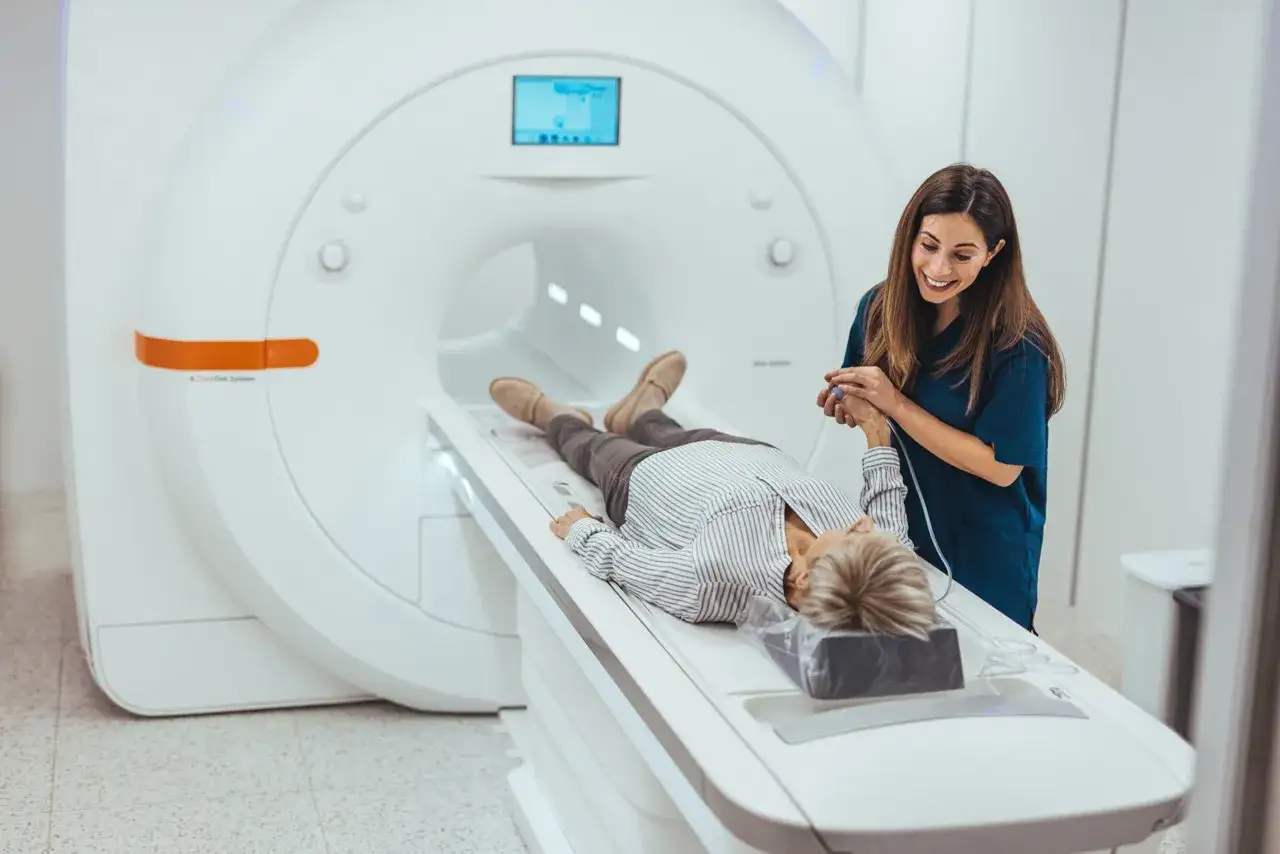 Eine Patientin liegt für eine MRT Untersuchung auf einer Liege, während eine freundliche Ärztin ihr die Hand hält.