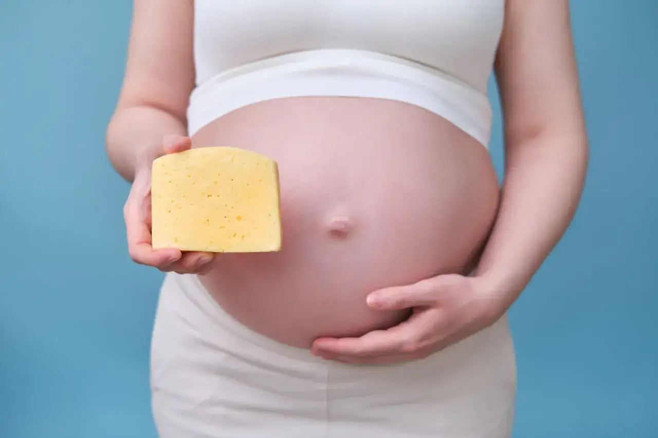 Schwangere hält ein Stück Käse neben ihrem Bauch. Vielleicht ein Hinweis auf die Ernährung während der Schwangerschaft, wie z.B. Mozzarella.