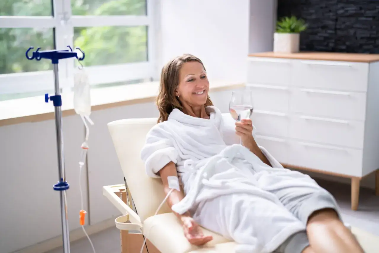 Frau genie&szlig;t eine Vitamin C Infusion, w&auml;hrend sie entspannt in einem Bademantel sitzt und ein Glas Wasser trinkt.