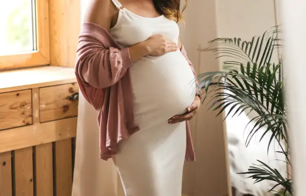 Frau in weißem Kleid und rosafarbener Strickjacke hält ihren Bauch, 17 ssw.