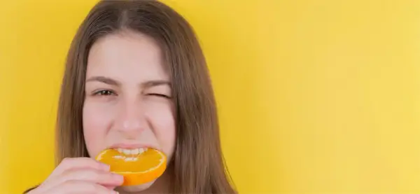 Junge Frau mit einem Stück Orange im Mund, die versucht, nach Weisheitszahn-OP wieder feste Nahrung zu essen.
