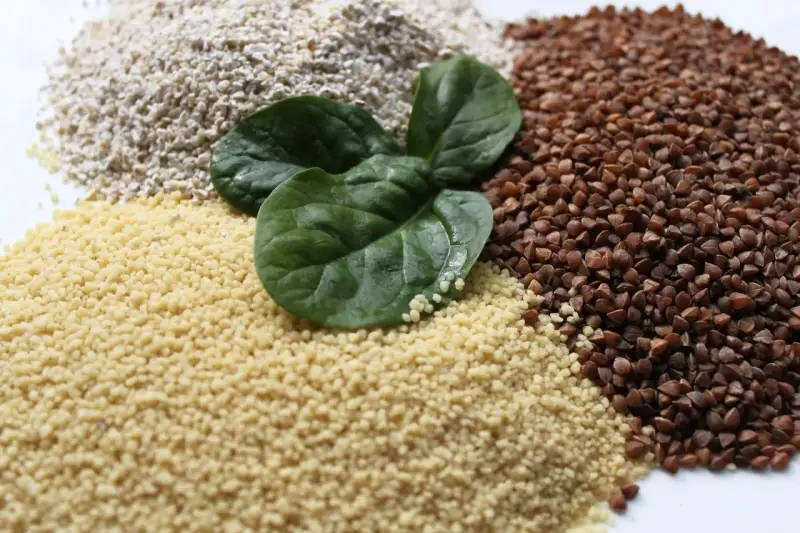 Haufen von Couscous, Buchweizen und Haferflocken, garniert mit frischen Spinatblättern. Ist Couscous gesund? Ja, es ist eine gute Quelle für Ballaststoffe.